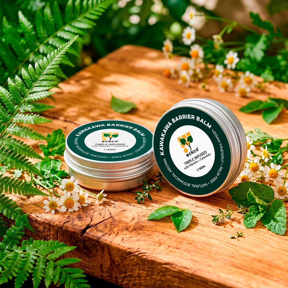 Two silver tins of eckco Kawakawa Barrier Balm resting on a rustic wooden board, surrounded by lush green fern fronds and small white chamomile flowers in bright, dappled sunlight