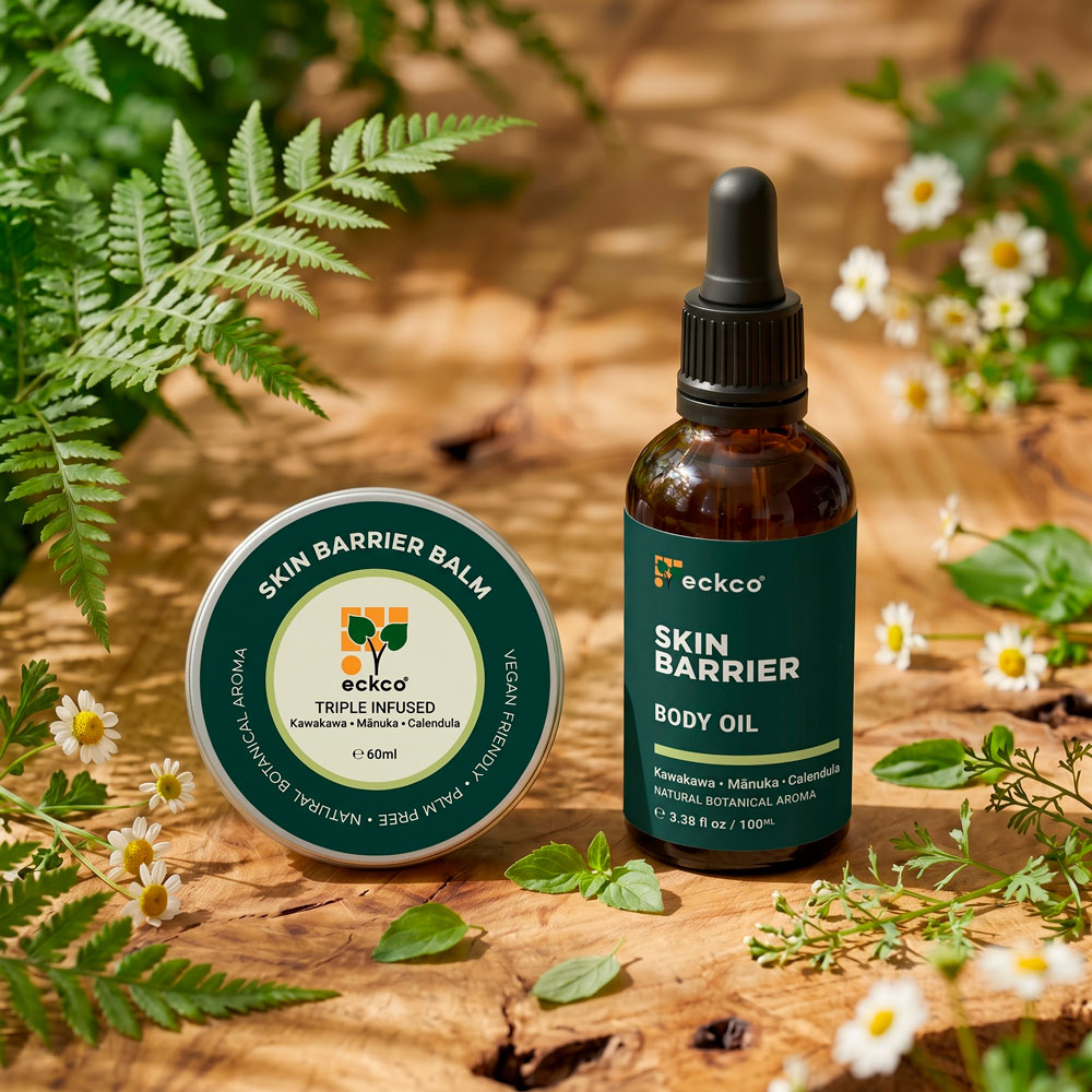 A top-down view of two eckco skincare products resting on a rustic wooden surface surrounded by fern fronds and small white flowers. On the left is a round metal tin of "Skin Barrier Balm" (60ml), and on the right is an amber glass dropper bottle of "Skin Barrier Body Oil" (100ml). Both product labels display green branding and note ingredients including Kawakawa, Mānuka, and Calendula