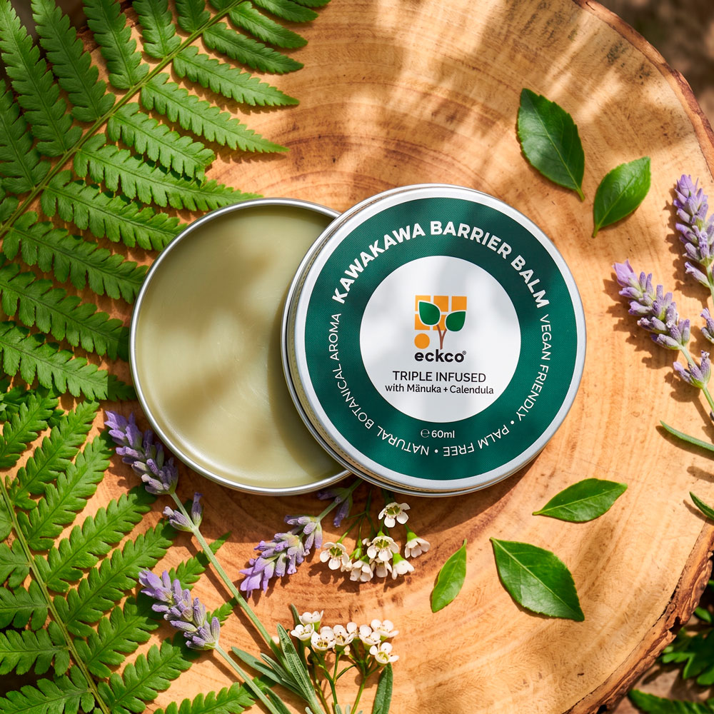 A tin of eckco Kawakawa Barrier Balm, open on a natural wood slice alongside its lid, decorated with fern fronds, lavender sprigs, and small white flowers. The lid displays the brand logo and labels reading "Kawakawa Barrier Balm", "Triple Infused with Mānuka + Calendula", "Palm Free", and "Vegan Friendly