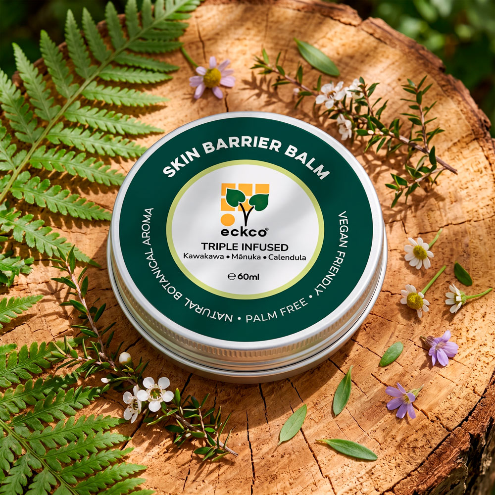A top-down view of a round 60ml tin of eckco Skin Barrier Balm resting on a rustic wooden log cross-section, surrounded by green ferns, small white mānuka flowers, and purple blossoms. The dark green and white label displays the eckco logo and reads "SKIN BARRIER BALM", "TRIPLE INFUSED Kawakawa • Mānuka • Calendula", "NATURAL BOTANICAL AROMA", "PALM FREE", and "VEGAN FRIENDLY