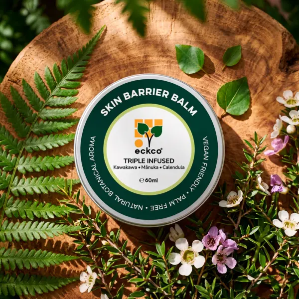 A top-down view of a tin of eckco Skin Barrier Balm resting on a rustic wood slice, surrounded by a green fern frond, scattered leaves, and blooming mānuka branches with white and pink flowers. The green and white label displays the brand name "eckco", "SKIN BARRIER BALM", "TRIPLE INFUSED", "Kawakawa • Mānuka • Calendula", and "e 60ml", encircled by text reading "NATURAL BOTANICAL AROMA • PALM FREE • VEGAN FRIENDLY