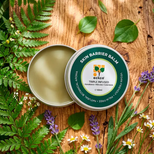 An overhead view of an open 60ml tin of eckco Skin Barrier Balm showing a smooth, light-green balm beside its dark green and white lid. The tin rests on a rustic wooden surface, framed by green fern leaves, heart-shaped leaves, purple lavender, and small white flowers