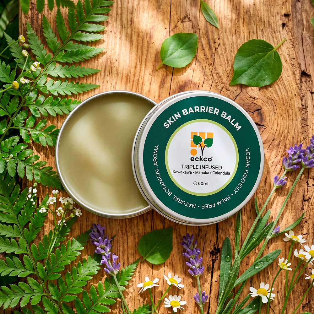 An overhead view of an open 60ml tin of eckco Skin Barrier Balm showing a smooth, light-green balm beside its dark green and white lid. The tin rests on a rustic wooden surface, framed by green fern leaves, heart-shaped leaves, purple lavender, and small white flowers