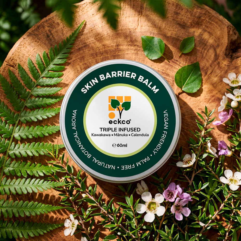 Top-down view of a 60ml tin of eckco Skin Barrier Balm resting on a rustic wooden slice, surrounded by fresh green fern fronds, kawakawa leaves, and flowering mānuka branches with small white and pink blossoms. The product label reads "SKIN BARRIER BALM", "eckco", and "TRIPLE INFUSED Kawakawa • Mānuka • Calendula