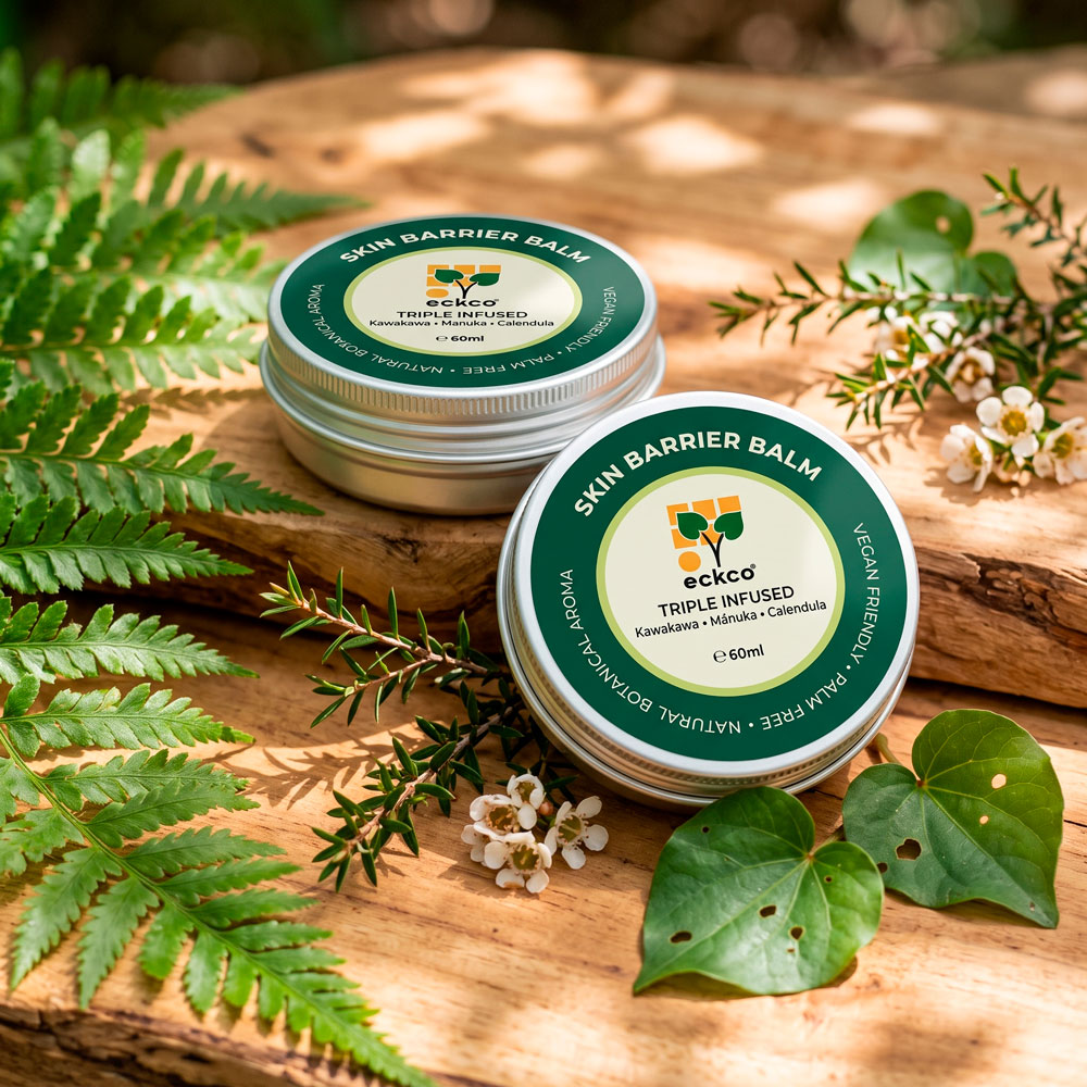 Two round silver tins of eckco Skin Barrier Balm, labeled "Triple Infused with Kawakawa, Mānuka, Calendula", displayed on a rustic wooden surface surrounded by lush fern fronds, small white Mānuka blossoms, and heart-shaped Kawakawa leaves in dappled sunlight