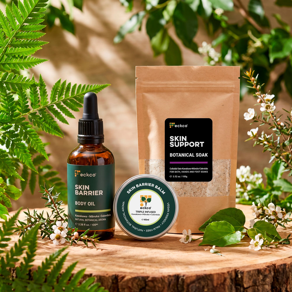 Three eckco skincare products arranged on a rustic wooden log slice, surrounded by green fern leaves, foliage, and small white mānuka blossoms in warm sunlight. From left to right: * An amber glass dropper bottle labelled "eckco SKIN BARRIER BODY OIL". * A round metal tin labelled "SKIN BARRIER BALM". * A brown kraft paper pouch with a black label reading "eckco SKIN SUPPORT BOTANICAL SOAK