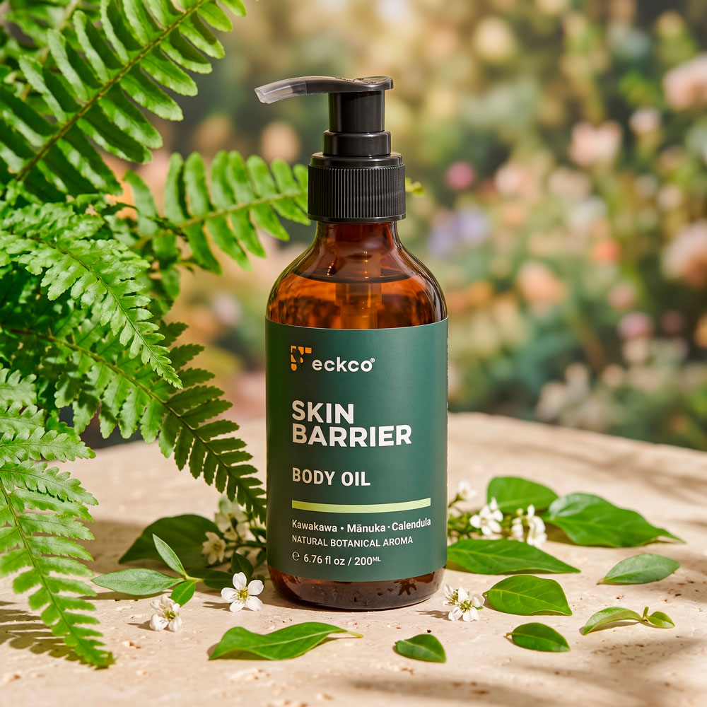 A 200mL amber glass pump bottle of eckco Skin Barrier Body Oil sitting upright on a light beige stone surface. The dark green label displays the brand name, product title, and key ingredients: Kawakawa, Mānuka, and Calendula. Lush green fern fronds frame the bottle on the left, while small white blossoms and green leaves lie scattered around its base against a soft-focus garden background