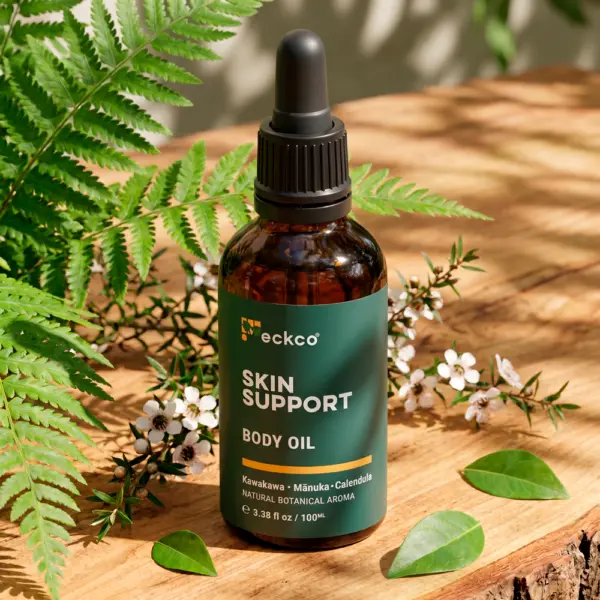An amber glass dropper bottle of Eckco Skin Support Body Oil with a dark green label listing Kawakawa, Mānuka, and Calendula. The bottle is set on a rustic wooden surface surrounded by lush green fern fronds and a flowering mānuka branch with small white blossoms in dappled sunlight