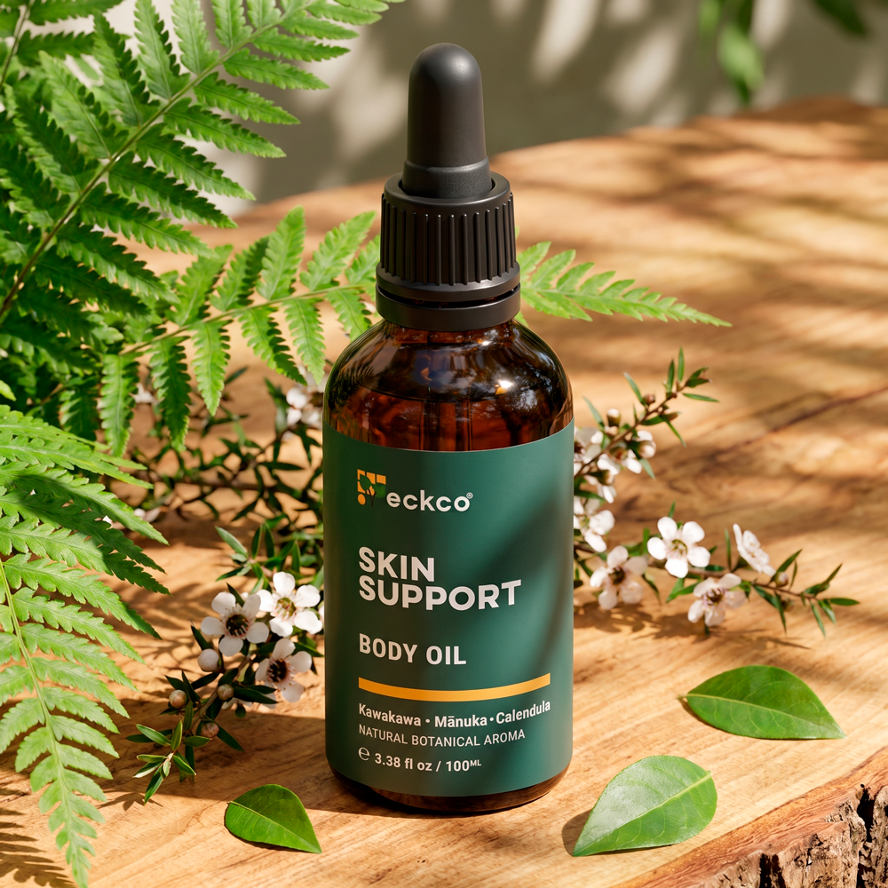 An amber glass bottle of eckco Skin Support Body Oil with a black dropper cap, resting on a wooden surface in dappled sunlight. The dark green label displays the text "eckco SKIN SUPPORT BODY OIL" above a yellow accent bar, listing key ingredients "Kawakawa • Mānuka • Calendula". The bottle is surrounded by fresh green fern fronds, small green leaves, and flowering mānuka branches with delicate white blossoms