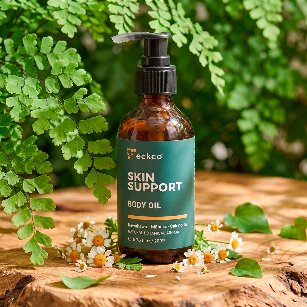 An amber glass pump bottle of eckco Skin Support Body Oil sitting on a rustic wooden surface. The teal label displays the brand name "eckco" and text reading "SKIN SUPPORT BODY OIL" along with ingredients "Kawakawa · Mānuka · Calendula". Small white and yellow chamomile flowers and green leaves are scattered around the base, with vibrant green fern foliage framing the scene