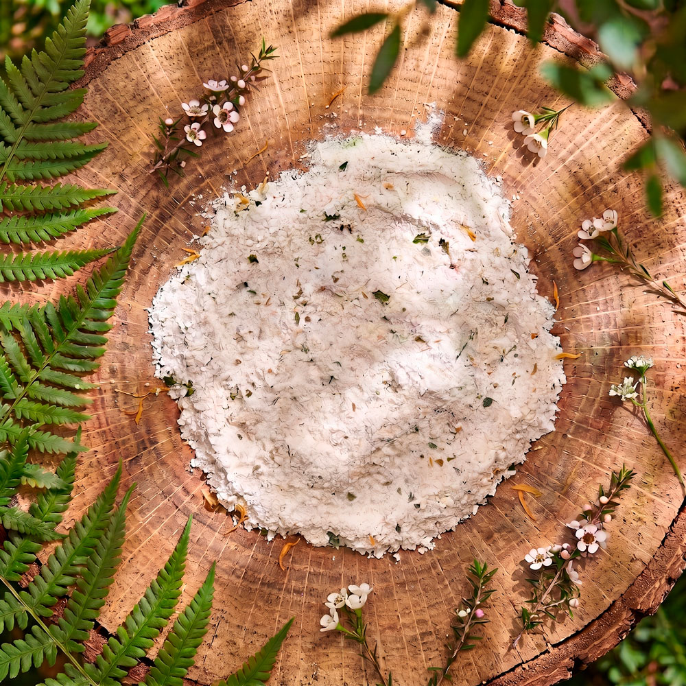 A top-down view of bath soak sitting in the centre of a round wooden log slice, surrounded by green fern fronds and small white and pink mānuka blossoms