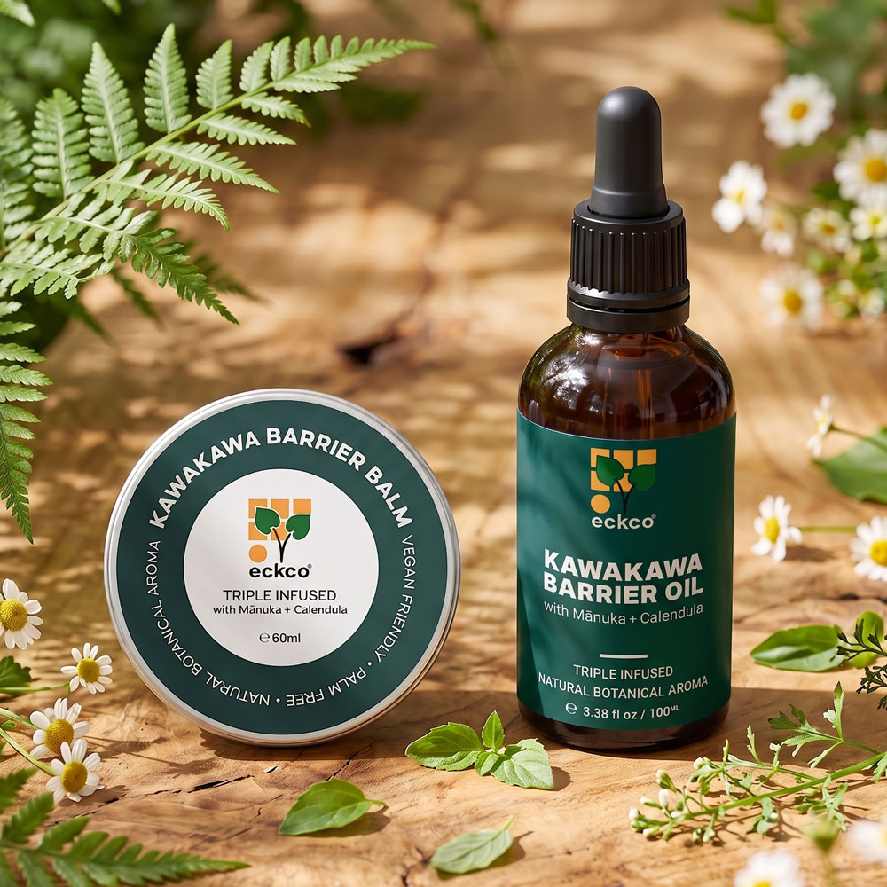 A round tin of eckco Kawakawa Barrier Balm and an amber glass bottle of eckco Kawakawa Barrier Oil with a dropper top, set on a wooden surface surrounded by green fern fronds and small white flowers in warm, natural sunlight