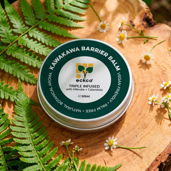 A top-down view of a 60ml tin of eckco Kawakawa Barrier Balm sitting on a slice of natural wood, surrounded by green fern fronds and white chamomile flowers. The tin lid features a dark green rim with text reading "KAWAKAWA BARRIER BALM" and a silver inner circle with the eckco logo and text stating "TRIPLE INFUSED with Mānuka + Calendula