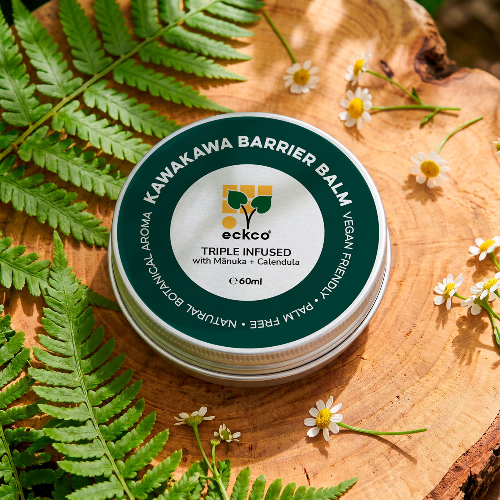 A top-down view of a 60ml tin of eckco Kawakawa Barrier Balm sitting on a slice of natural wood, surrounded by green fern fronds and white chamomile flowers. The tin lid features a dark green rim with text reading "KAWAKAWA BARRIER BALM" and a silver inner circle with the eckco logo and text stating "TRIPLE INFUSED with Mānuka + Calendula
