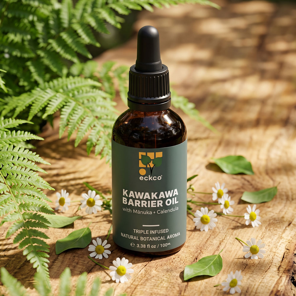 An amber glass dropper bottle of eckco Kawakawa Barrier Oil resting on a rustic wooden surface in dappled sunlight. The dark green label displays the product name "KAWAKAWA BARRIER OIL" with "Mānuka + Calendula". Surrounding the bottle on the wood are small white chamomile flowers, loose green leaves, and lush fern fronds extending from the left