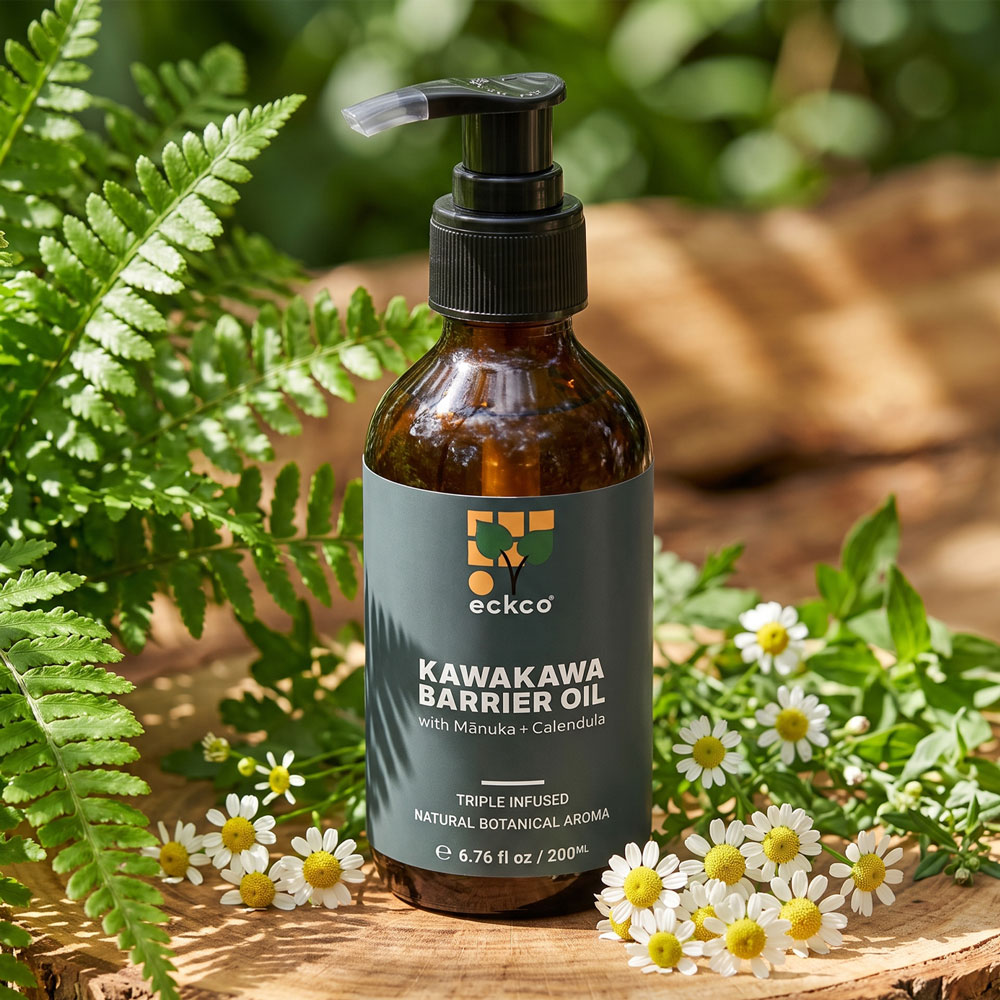 Alt text: A bottle of eckco Kawakawa Barrier Oil with Mānuka and Calendula sits on a wooden slice surrounded by green fern leaves and small white flowers
