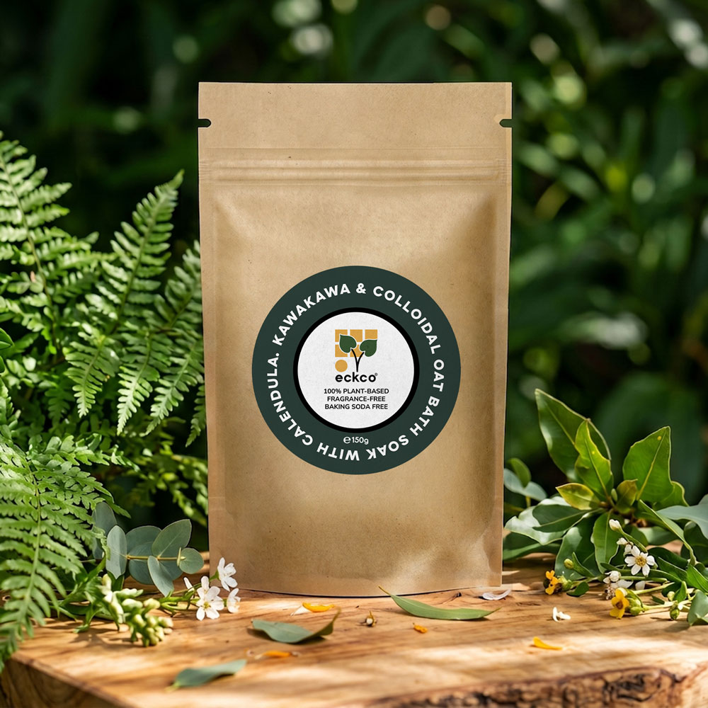 A brown kraft paper pouch of eckco bath soak with calendula, kawakawa, and colloidal oat, standing on a rustic wooden surface surrounded by lush green ferns and small white flowers in natural sunlight