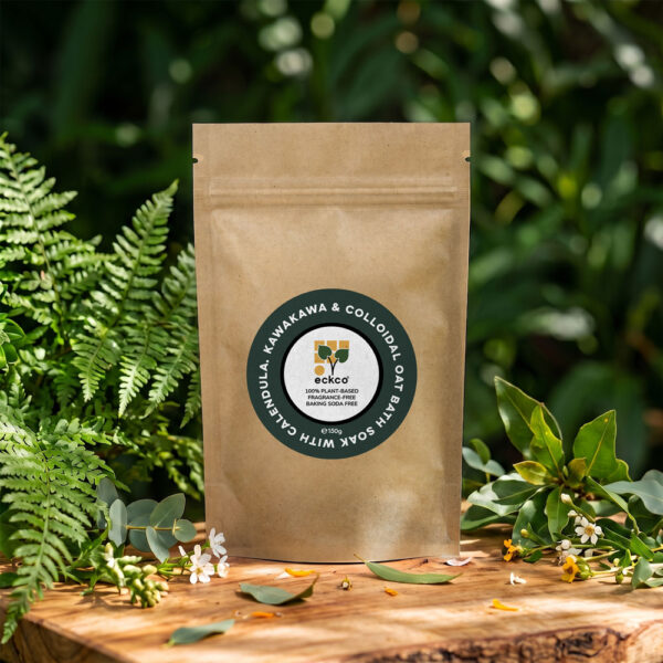 A brown kraft paper pouch bag of Eckco Bath Soak displayed on a rustic wooden surface amidst green ferns and foliage. The central circular dark green label reads "Bath Soak with Calendula, Kawakawa & Colloidal Oat" surrounding the brand logo "eckco