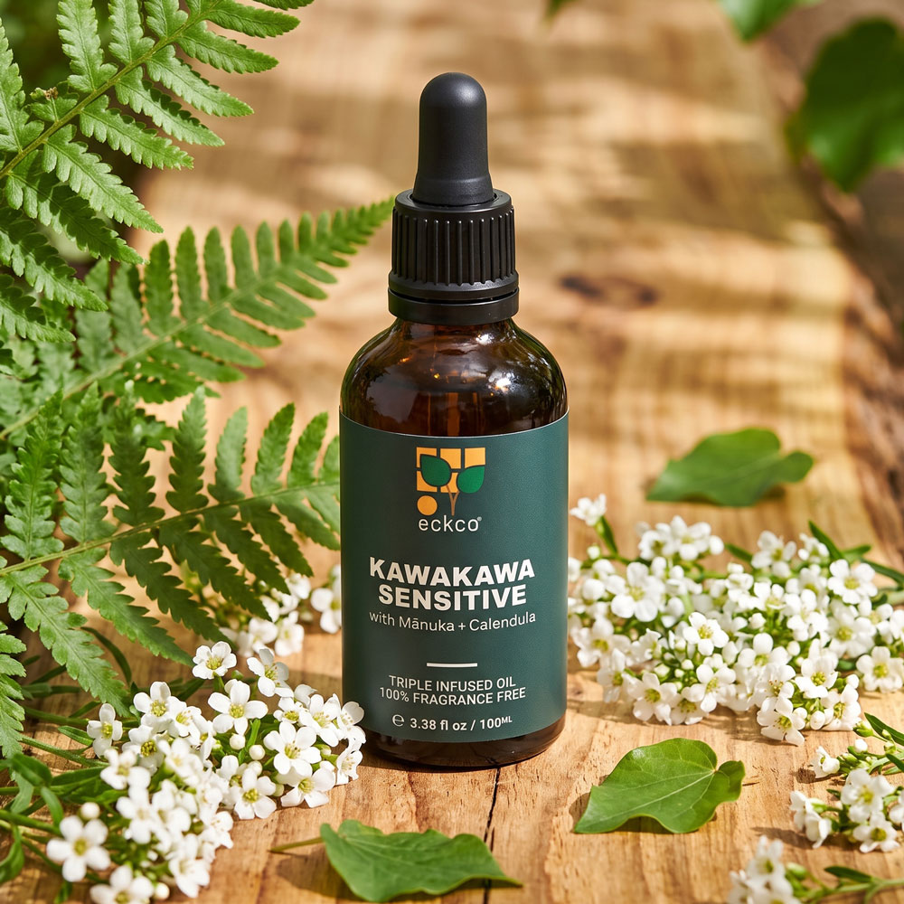 A 100ml amber glass dropper bottle of eckco Kawakawa Sensitive oil with Mānuka and Calendula, sitting on a rustic wooden surface surrounded by green fern fronds, small white flowers, and scattered kawakawa leaves