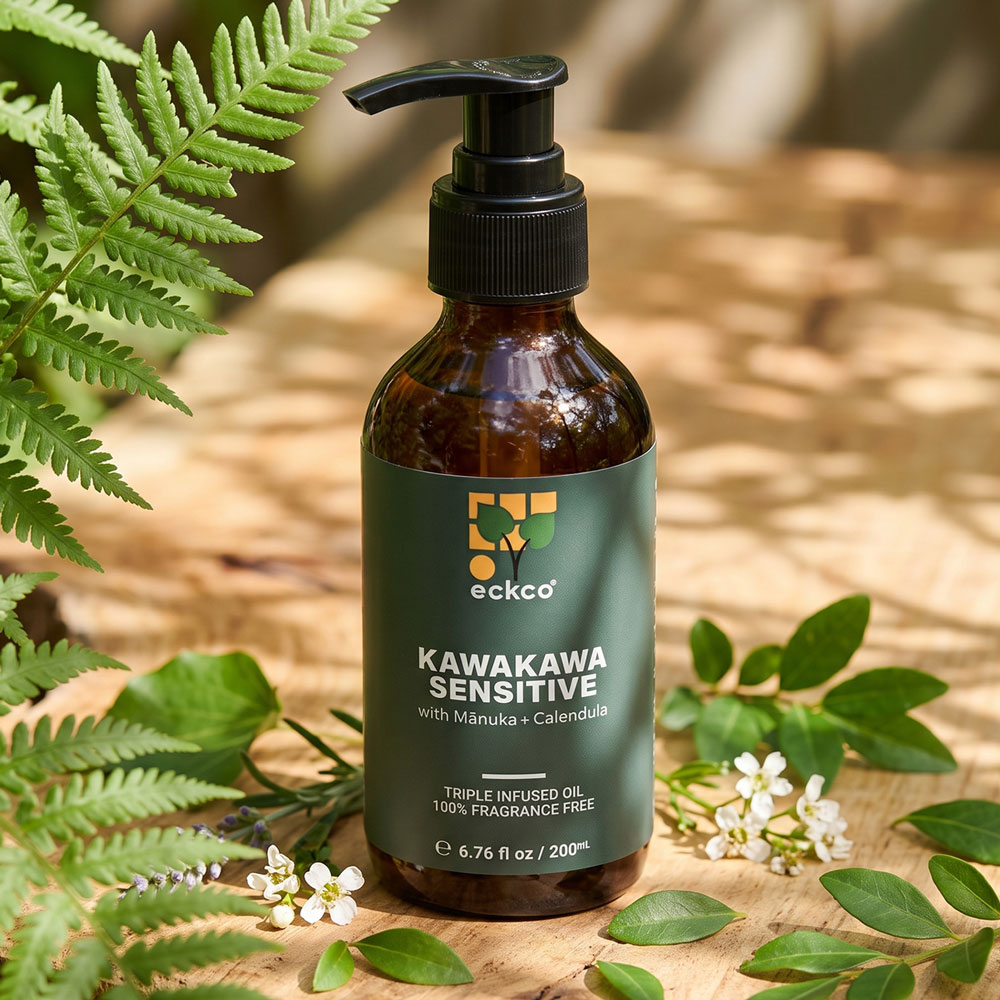 An amber glass bottle with a black pump dispenser labeled 'eckco KAWAKAWA SENSITIVE with Mānuka + Calendula'. The bottle sits on a light wooden surface surrounded by green fern fronds, sprigs of leaves, and small white flowers in dappled sunlight