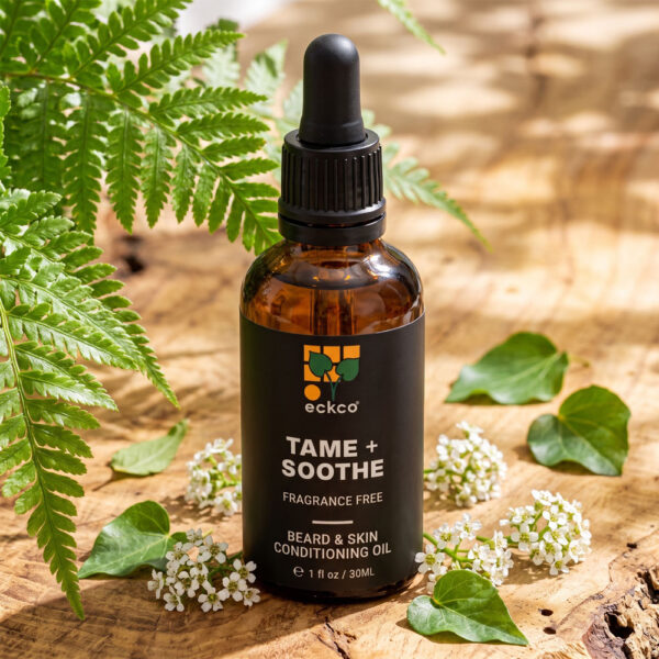 A dark amber glass dropper bottle with a black label reading "eckco TAME + SOOTHE FRAGRANCE FREE BEARD & SKIN CONDITIONING OIL" resting on a rustic wooden surface, framed by green fern leaves and small white flowers