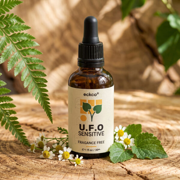 A brown glass dropper bottle labeled "eckco® U.F.O SENSITIVE FRAGANCE FREE" sitting upright on a rustic wooden slice. The bottle is surrounded by a green fern frond on the left and small white chamomile flowers with green leaves at its base, illuminated by warm natural sunlight
