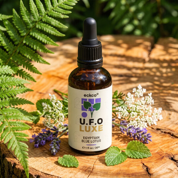 A brown glass dropper bottle with a white label reading "eckco U.F.O LUXE EGYPTIAN BLUE LOTUS" stands on a wooden stump, surrounded by green ferns, lavender sprigs, mint leaves, and delicate white flowers