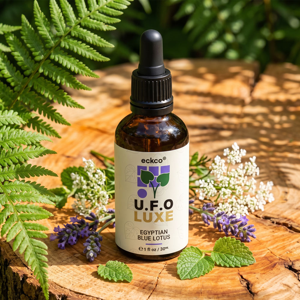 A brown glass dropper bottle with a white label reading "eckco U.F.O LUXE EGYPTIAN BLUE LOTUS" stands on a wooden stump, surrounded by green ferns, lavender sprigs, mint leaves, and delicate white flowers