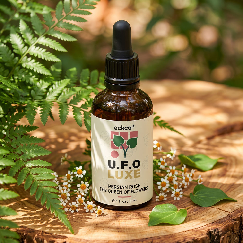 An amber glass bottle with a black dropper cap sitting upright on a cross-section of a wooden tree stump. The off-white label reads "eckco U.F.O LUXE PERSIAN ROSE THE QUEEN OF FLOWERS 1 fl oz / 30ML" alongside a green leaf graphic. Lush green fern fronds frame the left side of the bottle, while small white daisy-like flowers and green leaves are scattered around the base in warm, dappled sunlight
