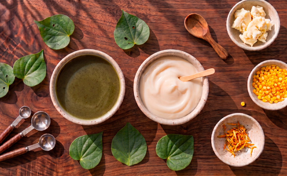 A top-down photograph of natural ingredients on a dark wooden surface. Centred are two large bowls, one containing a dark green liquid and the other a thick, cream-coloured paste. Several heart-shaped Kawakawa leaves are placed around the bowls. Three smaller bowls contain solid white chunks, small yellow pellets, and dried orange petals. The scene includes three metal measuring spoons with wooden handles and a wooden spoon