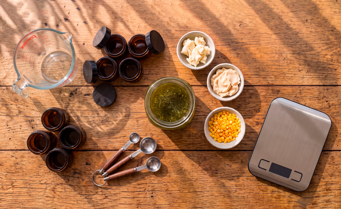 An overhead view of a wooden tabletop featuring a glass measuring jug, several small amber glass jars, and three small ceramic bowls. The bowls contain white solid chunks, a cream-coloured paste, and small yellow-orange pellets. A large glass jar in the centre contains a dark green liquid, commonly seen in Kawakawa infusions. Three metal measuring spoons with wooden handles and a digital kitchen scale are also present