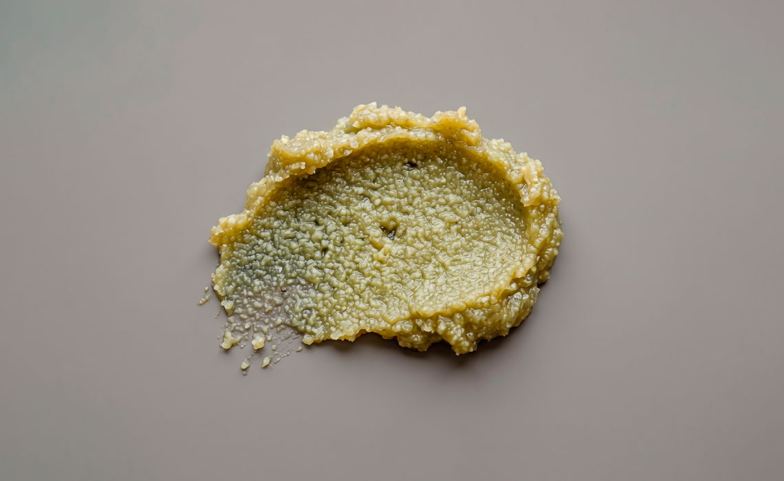 A flat lay macro view of a natural green kawakawa balm smear showing a grainy, bumpy texture caused by natural lipid crystallization.