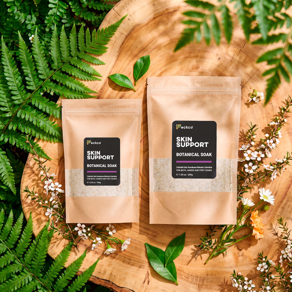 Flat lay of two brown kraft paper pouches of eckco Skin Support Botanical Soak sitting on a round wooden slab, framed by green ferns and white mānuka blossoms. The pouches come in 100g and 200g sizes with black labels displaying the product details