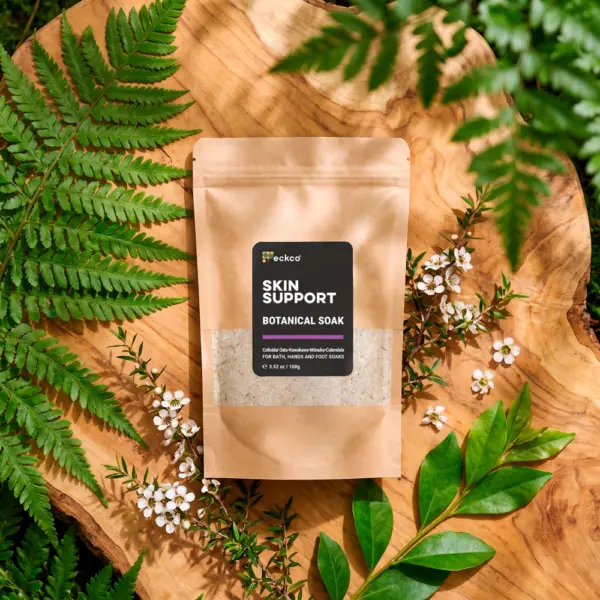 A top-down view of a brown kraft paper pouch of eckco Skin Support Botanical Soak resting on a wooden slab, surrounded by green fern fronds and mānuka branches with small white flowers