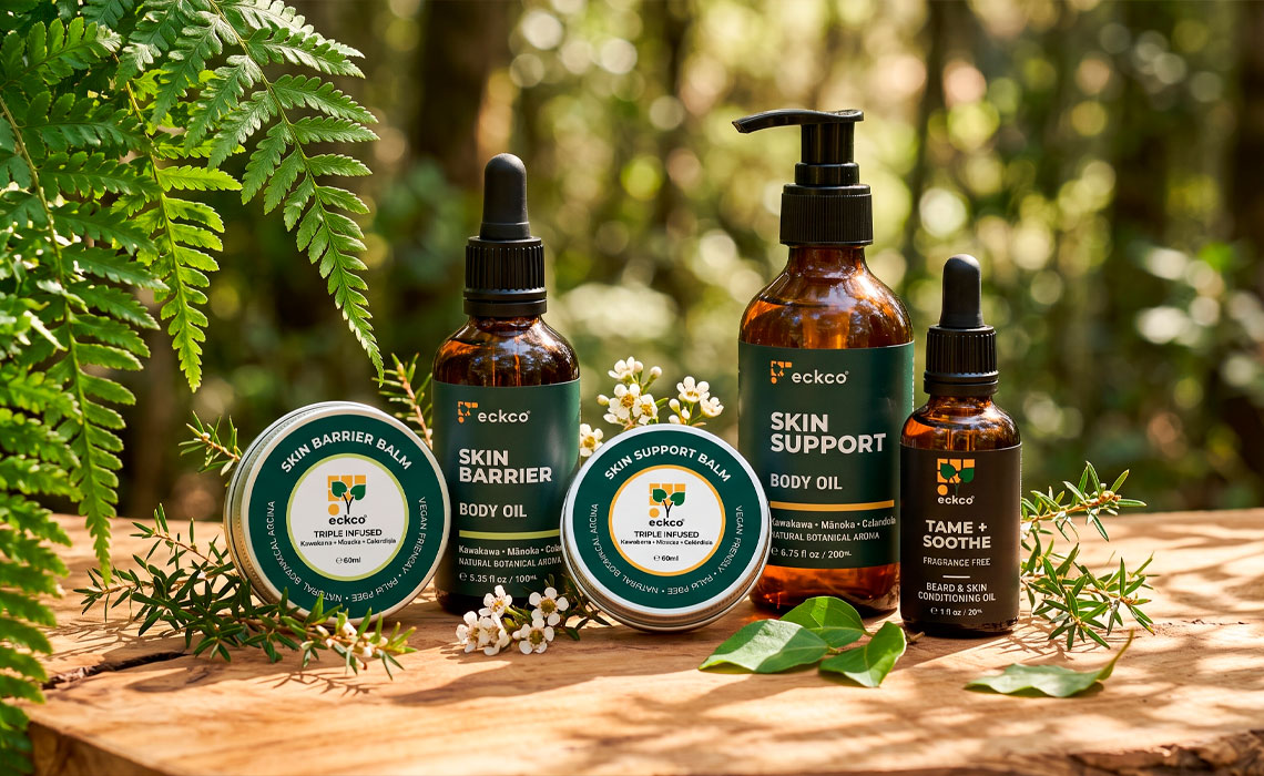 A selection of five eckco botanical skincare products arranged on a wooden surface against a sunlit, blurred forest background with green ferns and tiny white flowers. The amber glass bottles and silver tins include Skin Barrier Balm, Skin Barrier Body Oil, Skin Support Balm, Skin Support Body Oil, and Tame + Soothe Beard & Skin Conditioning Oil