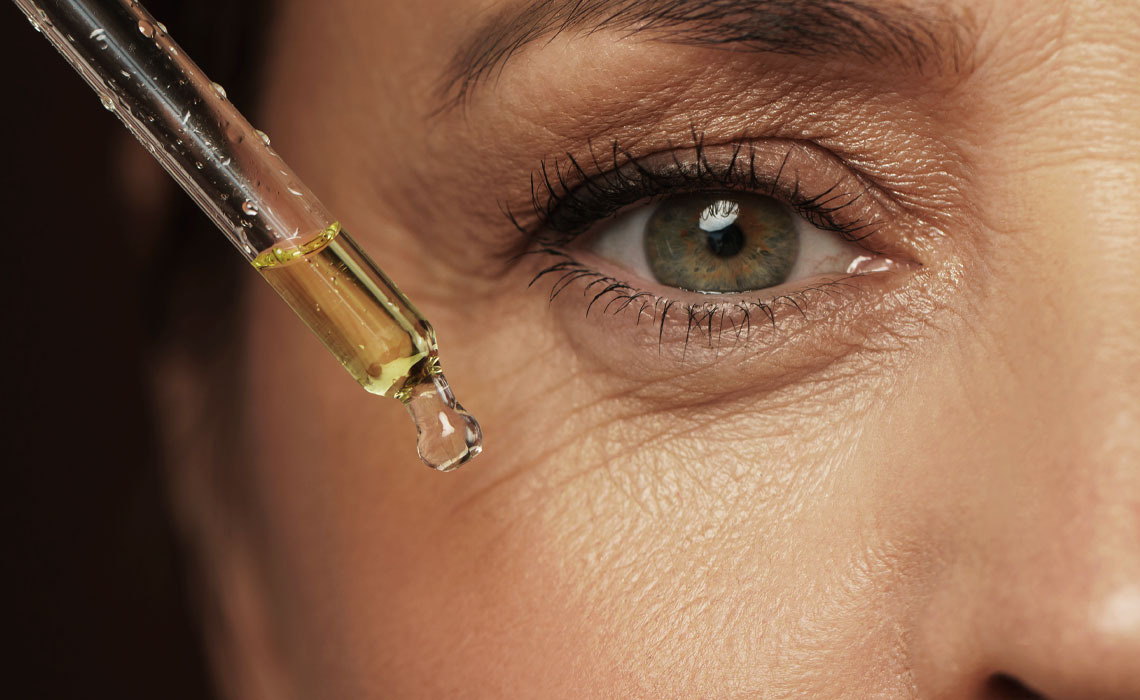 A close-up of mature skin around a person's green eye, with a glass dropper holding golden facial serum positioned near the cheek