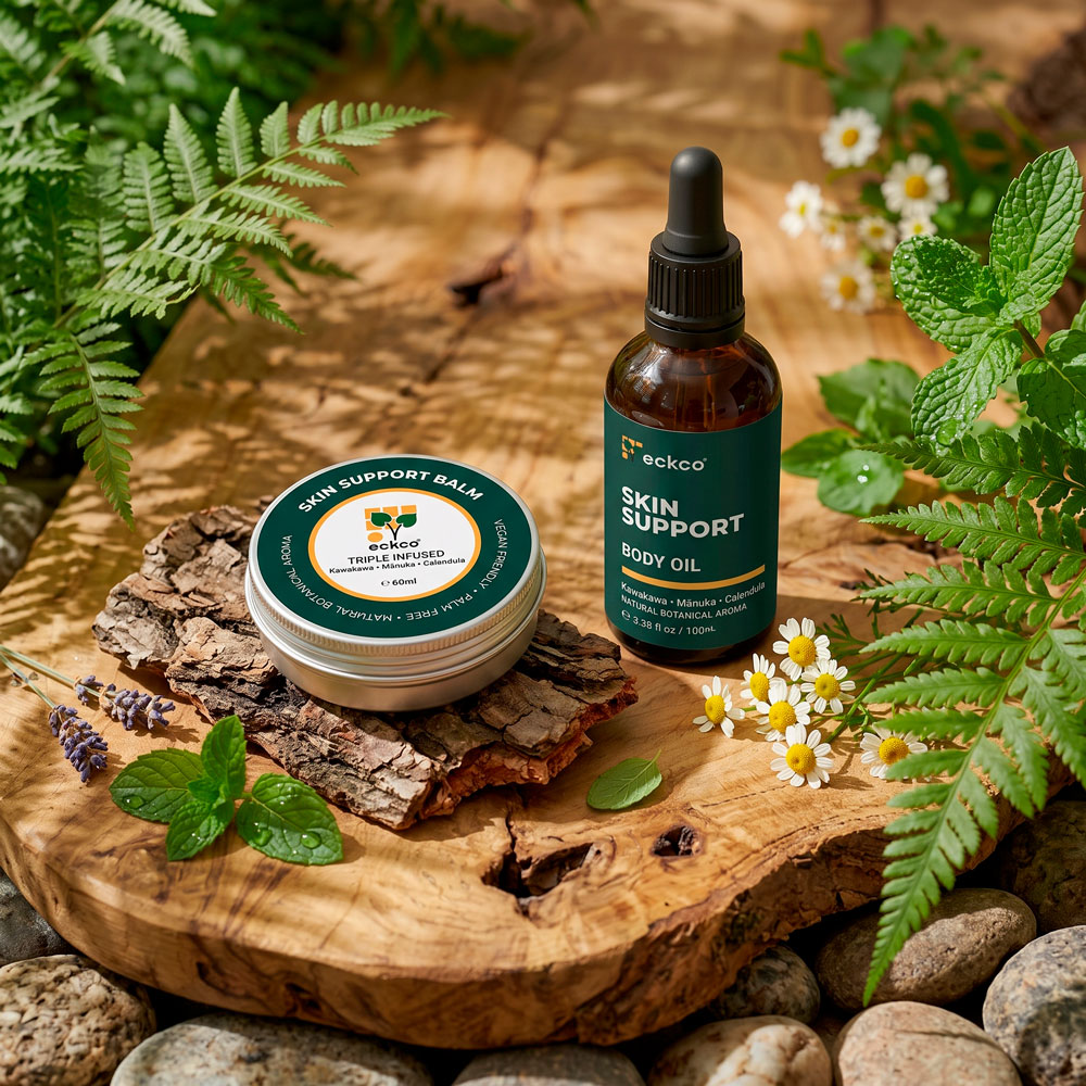 A metal tin of eckco Skin Support Balm resting on tree bark alongside an amber glass dropper bottle of eckco Skin Support Body Oil, arranged on a rustic wooden slab surrounded by green ferns, mint leaves, lavender, and small white flowers
