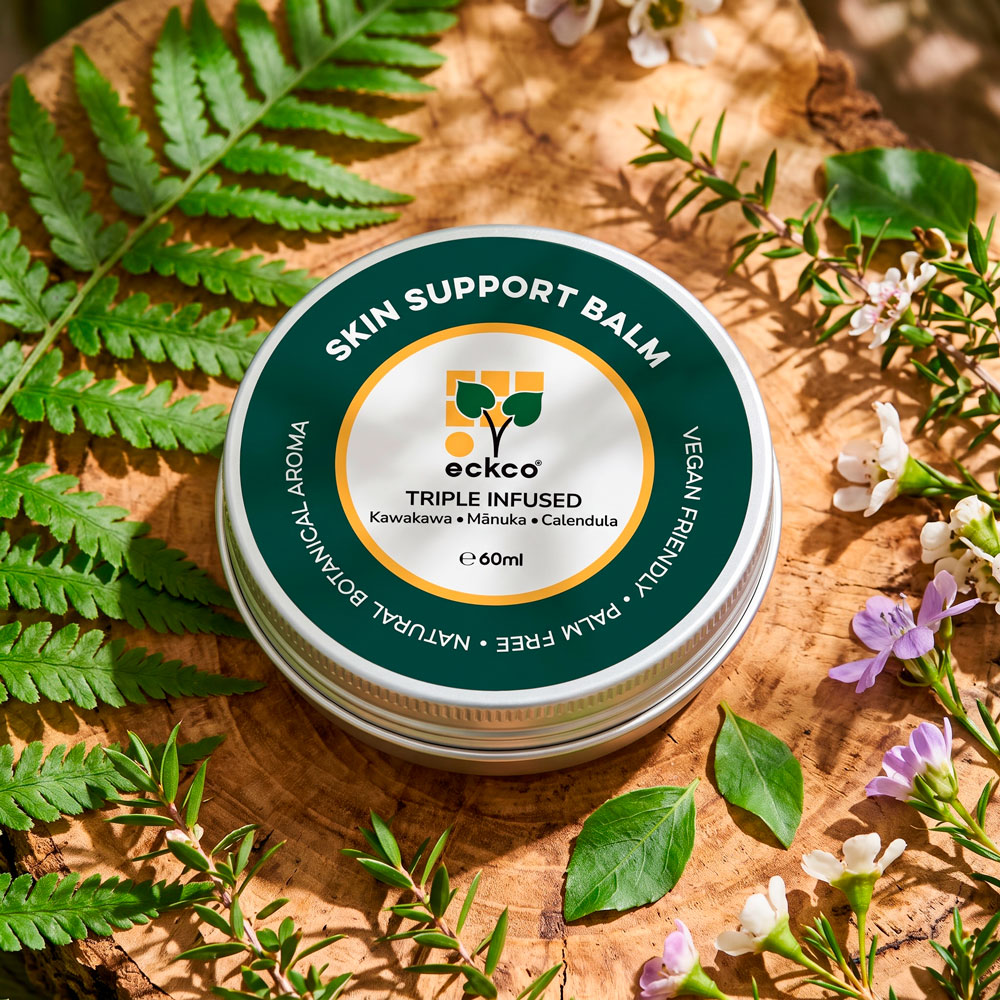 A top-down view of a 60ml tin of Eckco Skin Support Balm resting on a wooden log slice, surrounded by green fern leaves and small mānuka flowers in warm sunlight. The round green and white label reads "SKIN SUPPORT BALM", "eckco", "TRIPLE INFUSED", and "Kawakawa • Mānuka • Calendula