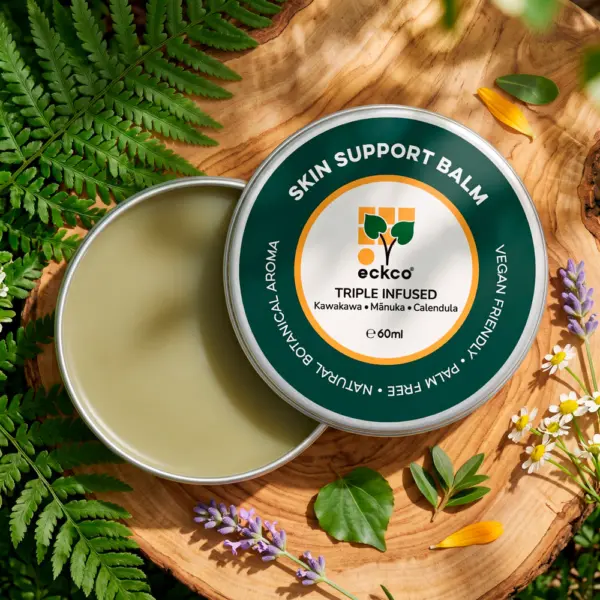 An overhead shot of an open 60ml silver tin of eckco Skin Support Balm resting on a rustic wood slice, surrounded by green fern fronds, sprigs of lavender, and small white chamomile flowers. Beside the smooth, light green balm, the dark green lid displays text reading "Skin Support Balm" and "Triple Infused: Kawakawa • Mānuka • Calendula