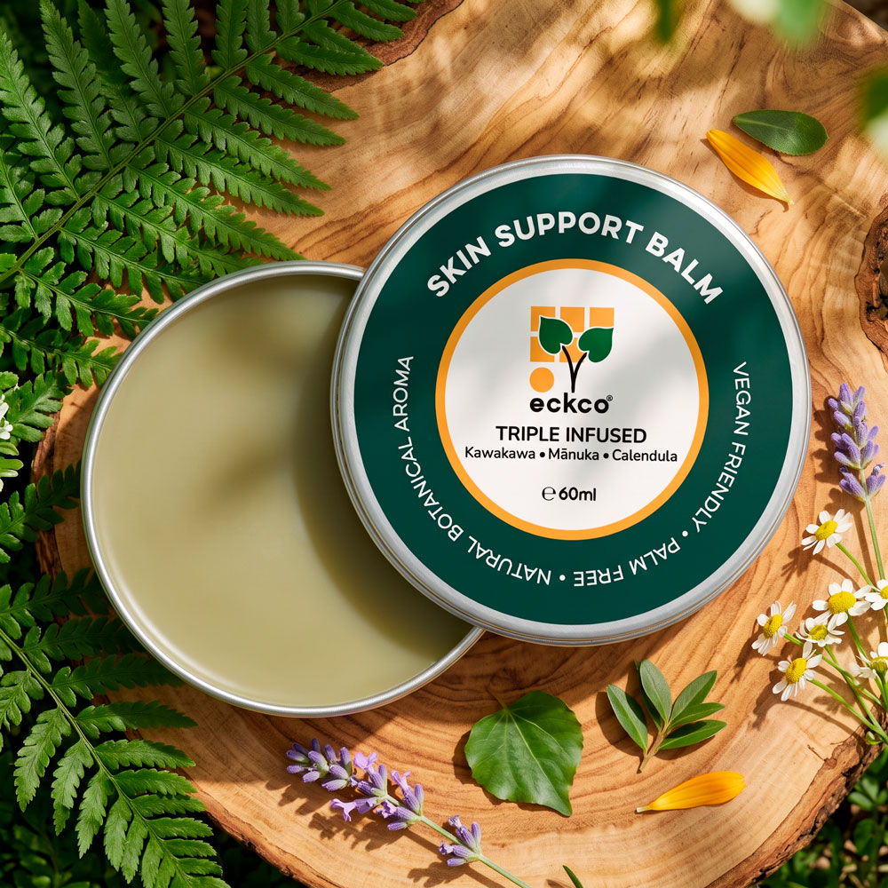 A high-angle, close-up shot of an open circular tin of eckco Skin Support Balm resting on a rustic wooden slice, surrounded by green fern fronds, lavender, and small chamomile flowers. The offset lid reveals a smooth, light-green balm inside. The lid features a dark green ring with white text reading "SKIN SUPPORT BALM" and a central white label displaying the eckco logo, "TRIPLE INFUSED", "Kawakawa • Mānuka • Calendula", and "e 60ml
