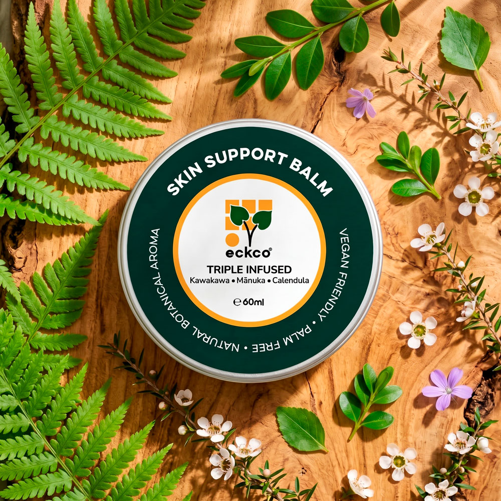 A top-down view of a round 60ml silver tin of eckco Skin Support Balm set on a textured wood grain background. The tin has a dark green circular label with white text reading "SKIN SUPPORT BALM" around the top and "NATURAL BOTANICAL AROMA • PALM FREE • VEGAN FRIENDLY" around the bottom. In the center, a white circle trimmed in orange showcases the eckco plant logo and text that reads "TRIPLE INFUSED Kawakawa • Mānuka • Calendula" above "e 60ml". The tin is surrounded by fresh green fern fronds, mānuka sprigs with small white flowers, and delicate purple blossoms