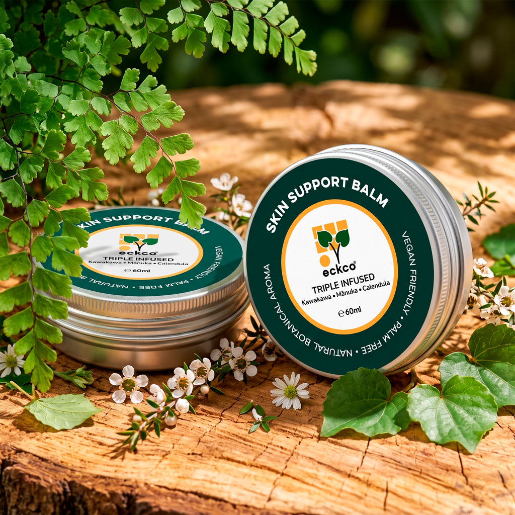 A photo of two silver tins of eckco Skin Support Balm displayed outdoors on a rustic wooden log slab, framed by green fern fronds and small white mānuka flowers. One tin stands upright in the foreground, clearly showing its dark green label with white and orange text that reads "SKIN SUPPORT BALM", "eckco", and "TRIPLE INFUSED Kawakawa • Mānuka • Calendula". A second tin rests horizontally beside it to the left