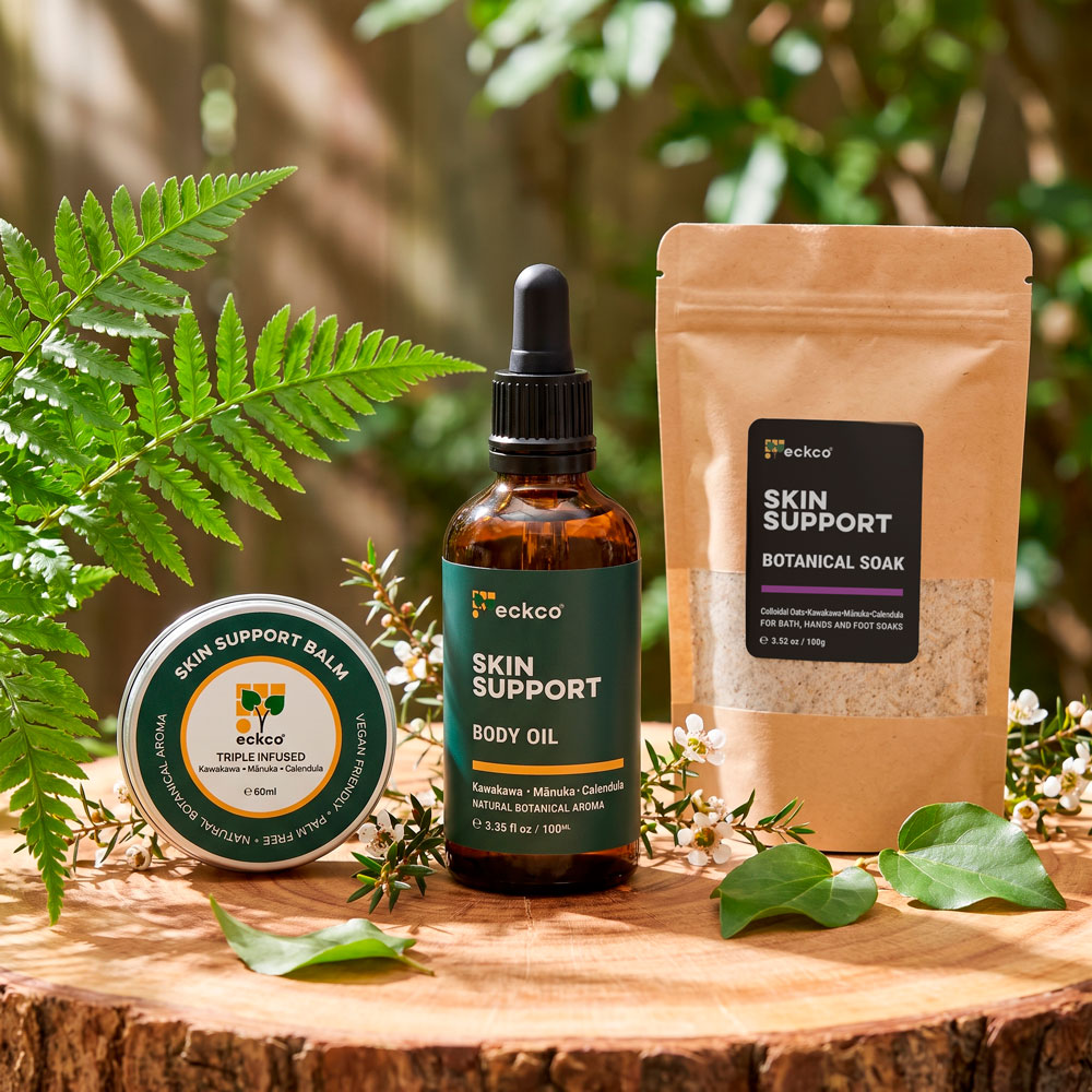 Three eckco Skin Support natural skincare products displayed on a slice of a wooden log amidst green ferns and botanical foliage in bright sunlight. The product range includes a round tin of Skin Support Balm on the left, an amber glass dropper bottle of Skin Support Body Oil in the centre, and a kraft paper pouch of Skin Support Botanical Soak on the right